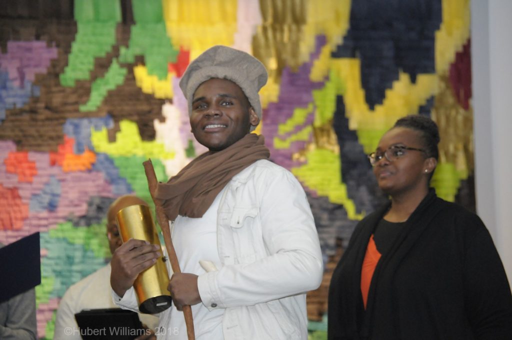 Photo of a man wearing brown scarf, holding stick and golden tumbler and woman wearing a glasses and black jacket at his back